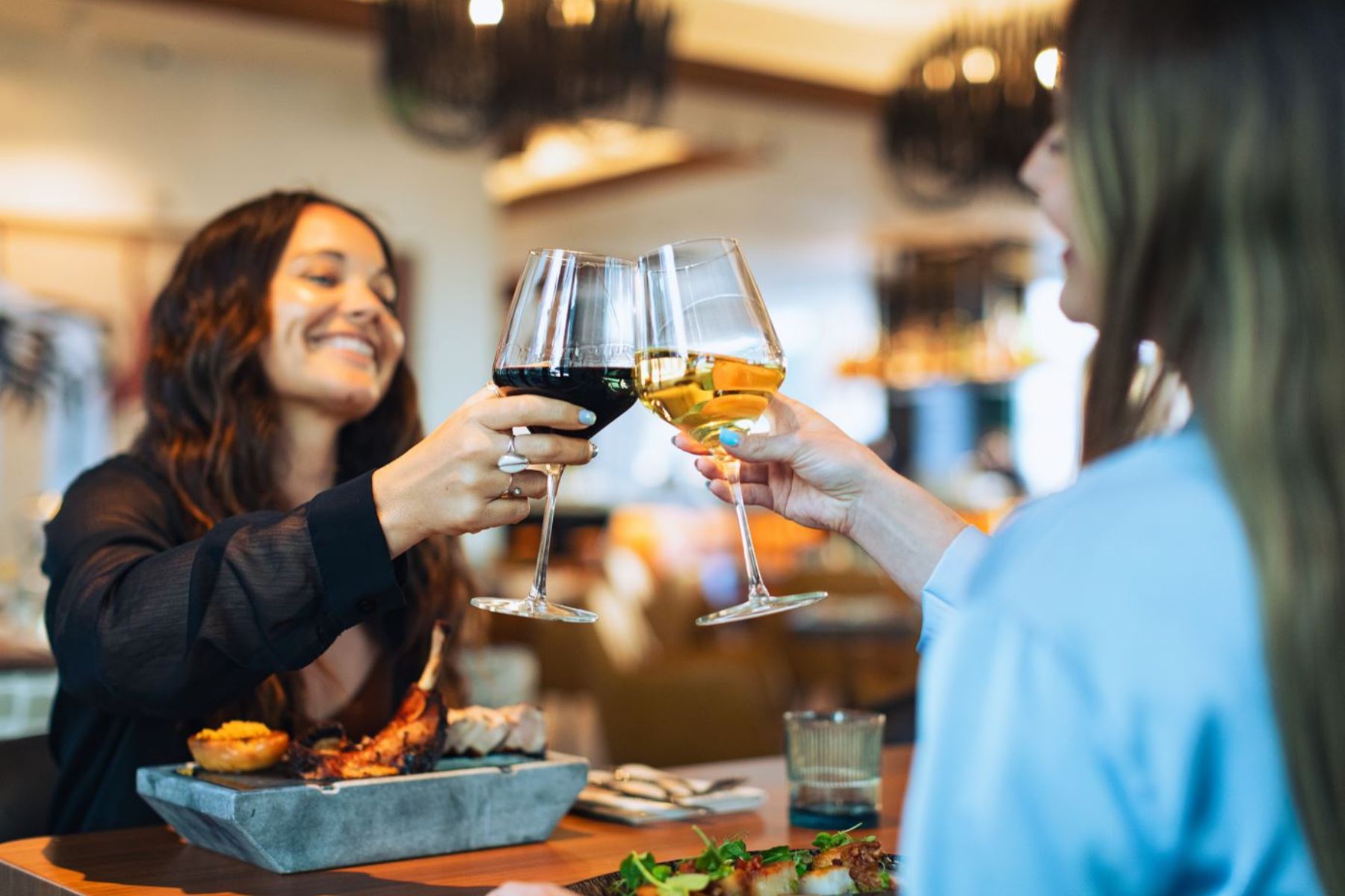 Two women sitting at a restaurant table clink wine glasses, one with red and one with white, while smiling and enjoying a meal—perfect for a festive Holiday Private Dining experience.
