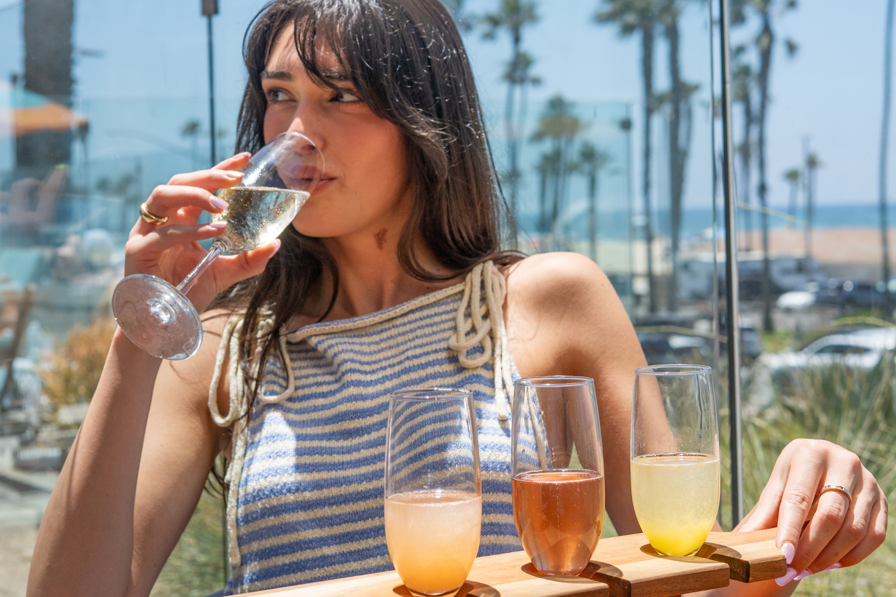 A woman in a striped top enjoys a Hangover Brunch, sipping her drink and holding a tray of assorted beverages, seated outdoors with palm trees and a beach in the background.