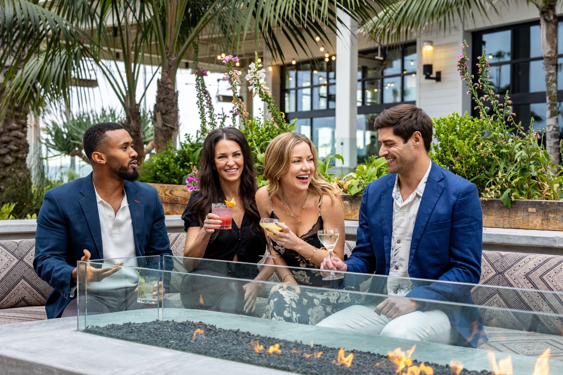 Four adults sit around an outdoor fire pit, smiling and holding drinks, as they enjoy cool culinary delights at Lōrea, with palm trees and lush plants in the background.