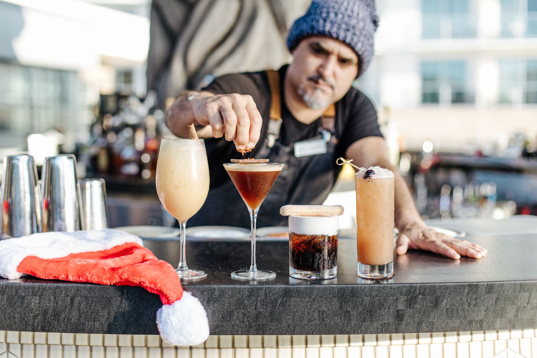 A bartender places a garnish on a cocktail beside three other drinks on a bar counter, with a Santa hat in the foreground, evoking the cozy spirit of Home for the Holidays.