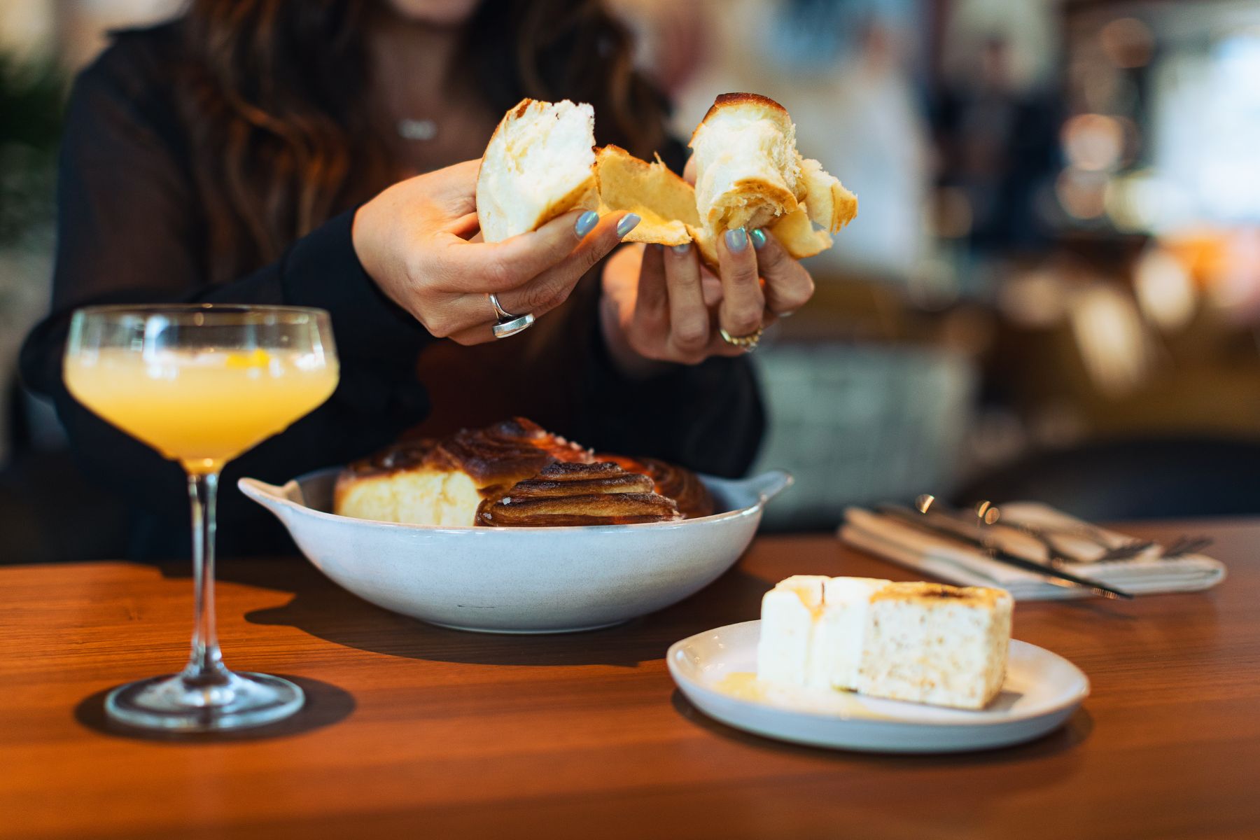 A person breaks apart a piece of bread at a restaurant table, enjoying Christmas dining with a bowl of bread, a plate piled high with more bread, and a glass of orange beverage in front of them.