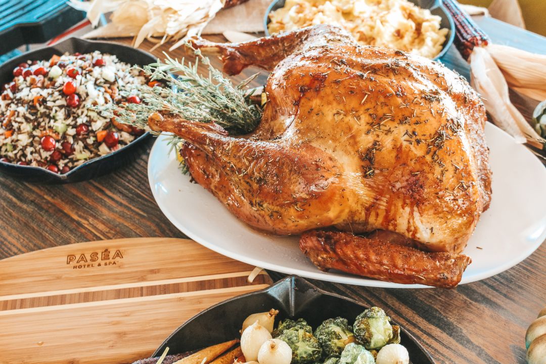 A whole roasted turkey garnished with herbs is served on a white platter, surrounded by classic Thanksgiving dinner sides like salad, Brussels sprouts, and mashed potatoes on a wooden table.