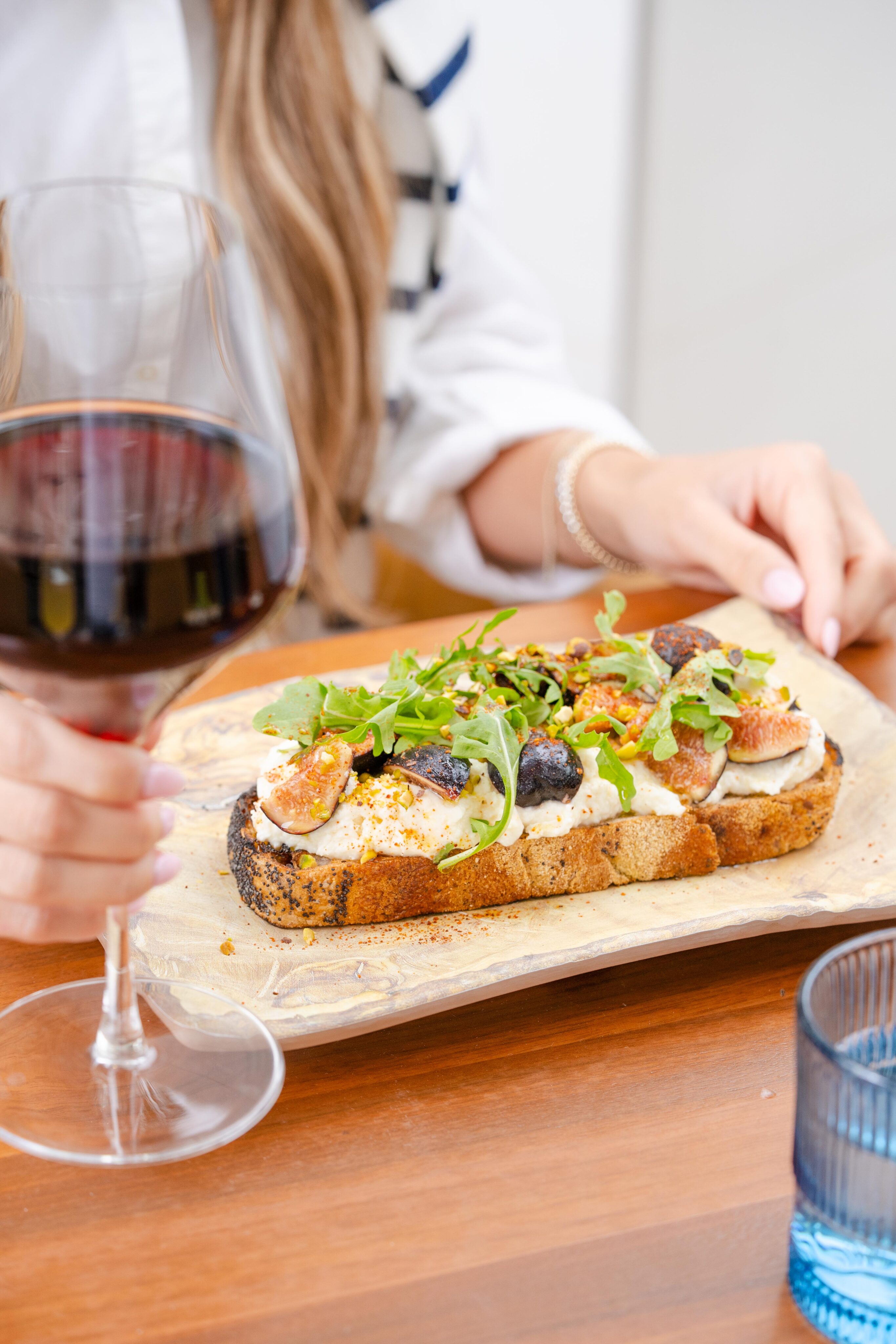A person holding a glass of red wine sits at a table with a slice of bread topped with figs, arugula, and cheese on a wooden board during the limited menu hours of 3-5PM.