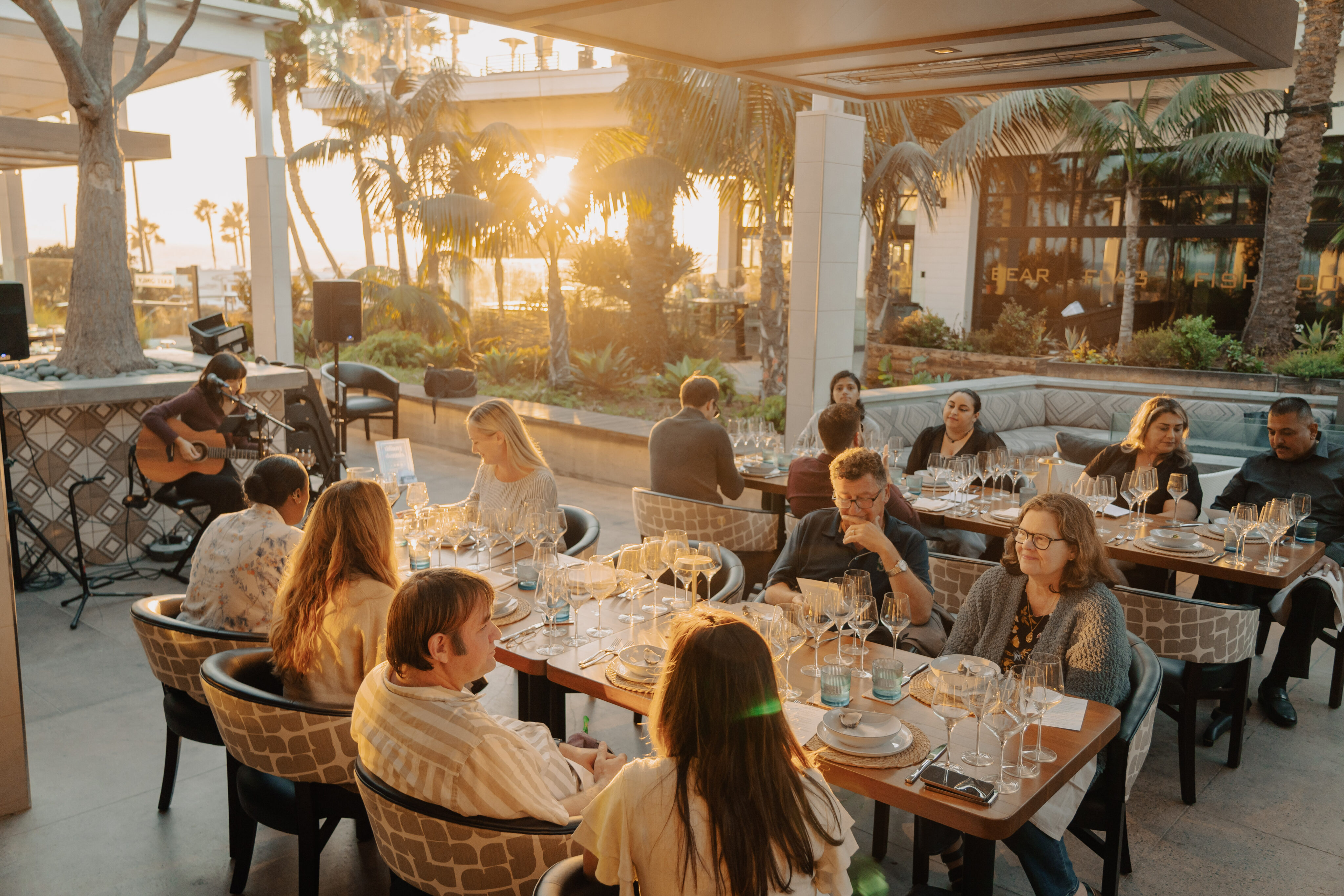 Guests enjoy the Taste of the Table Dinner Series at sunset, dining outdoors as a musician plays guitar beneath swaying palm trees, with Trinitas Cellars wines setting the perfect mood.