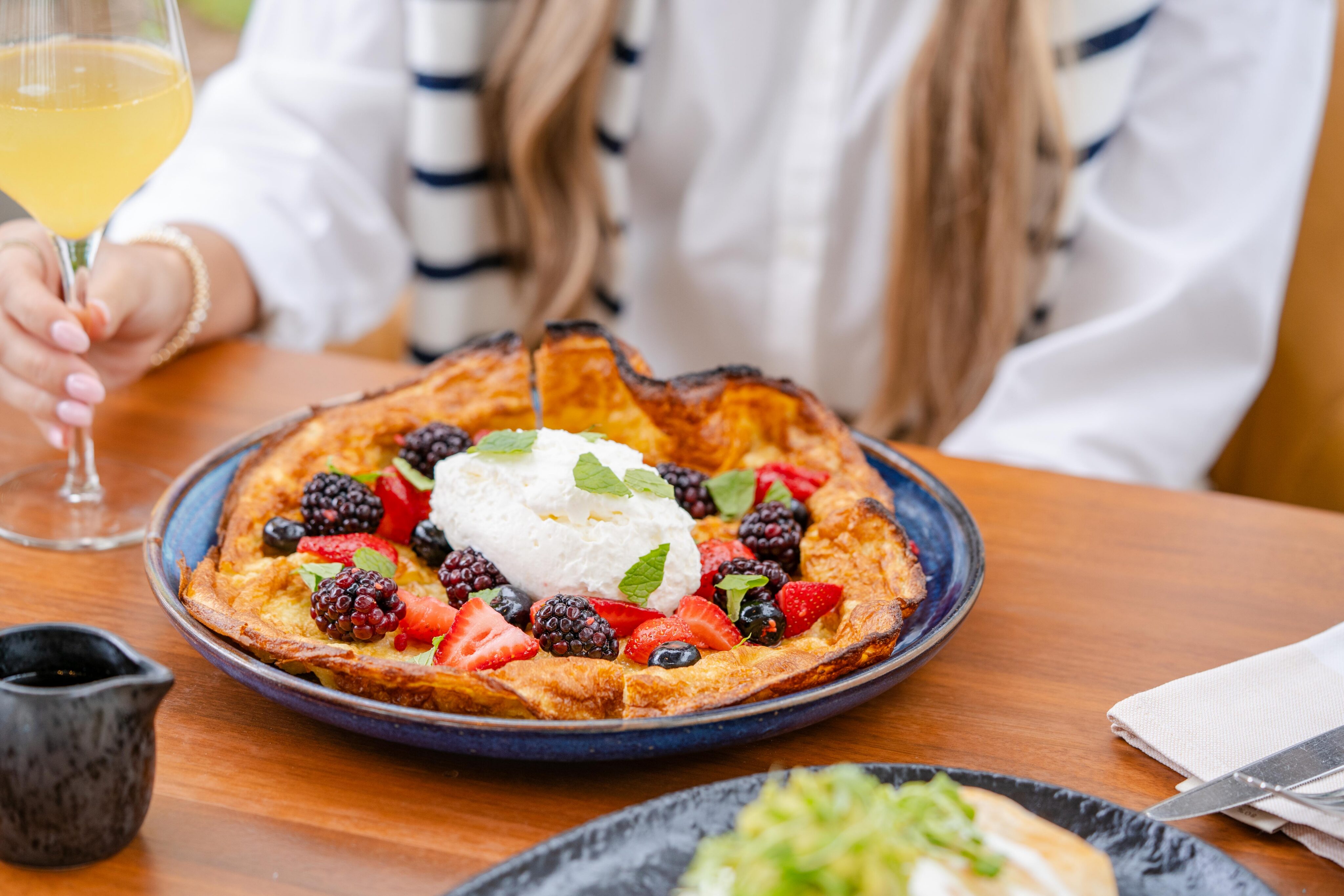 A plate with a large Dutch baby pancake topped with whipped cream, fresh strawberries, blackberries, and mint sits on a wooden table next to a person holding a glass of yellow beverage.
