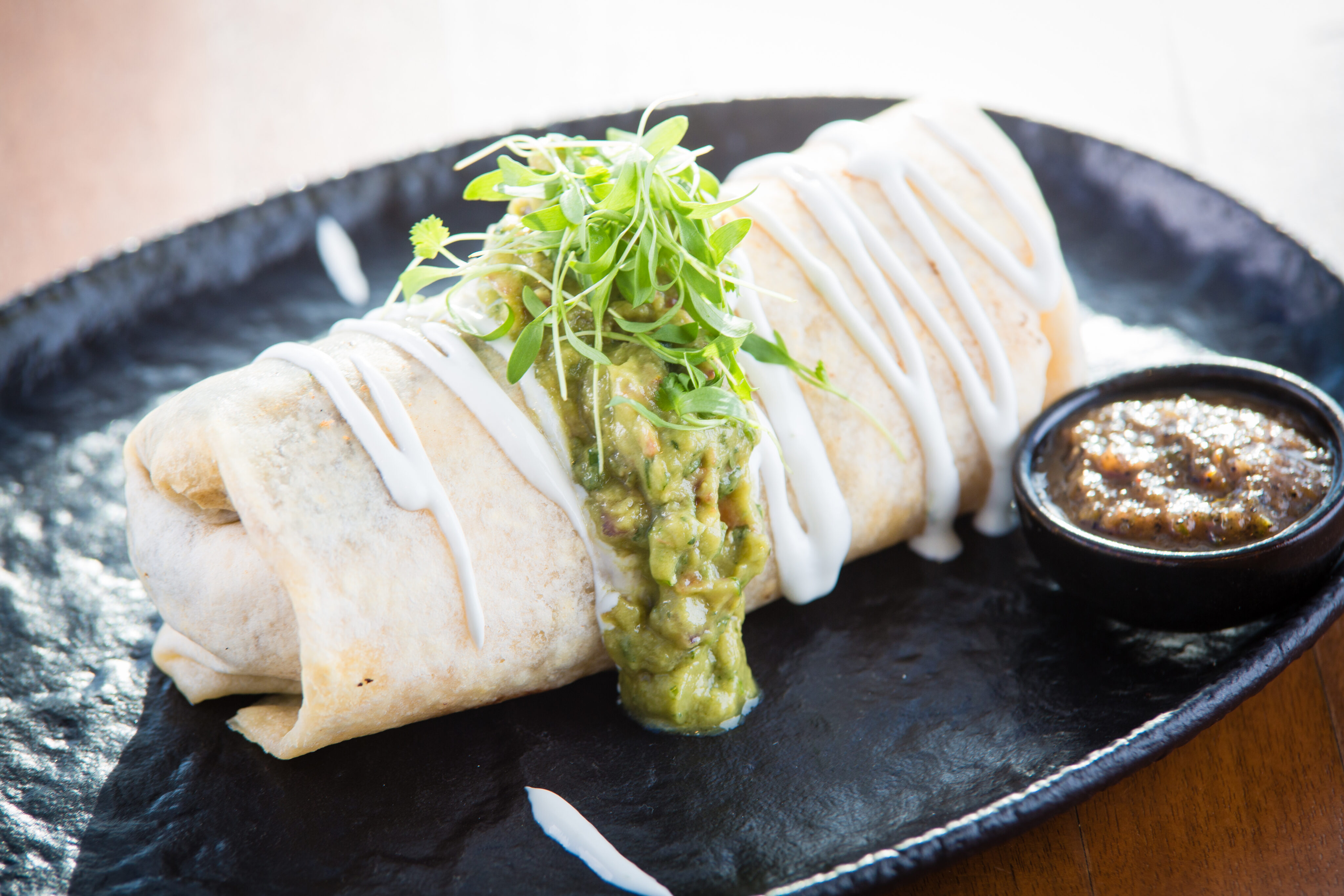A burrito topped with sour cream, guacamole, and microgreens is served on a black plate with a side of salsa.
