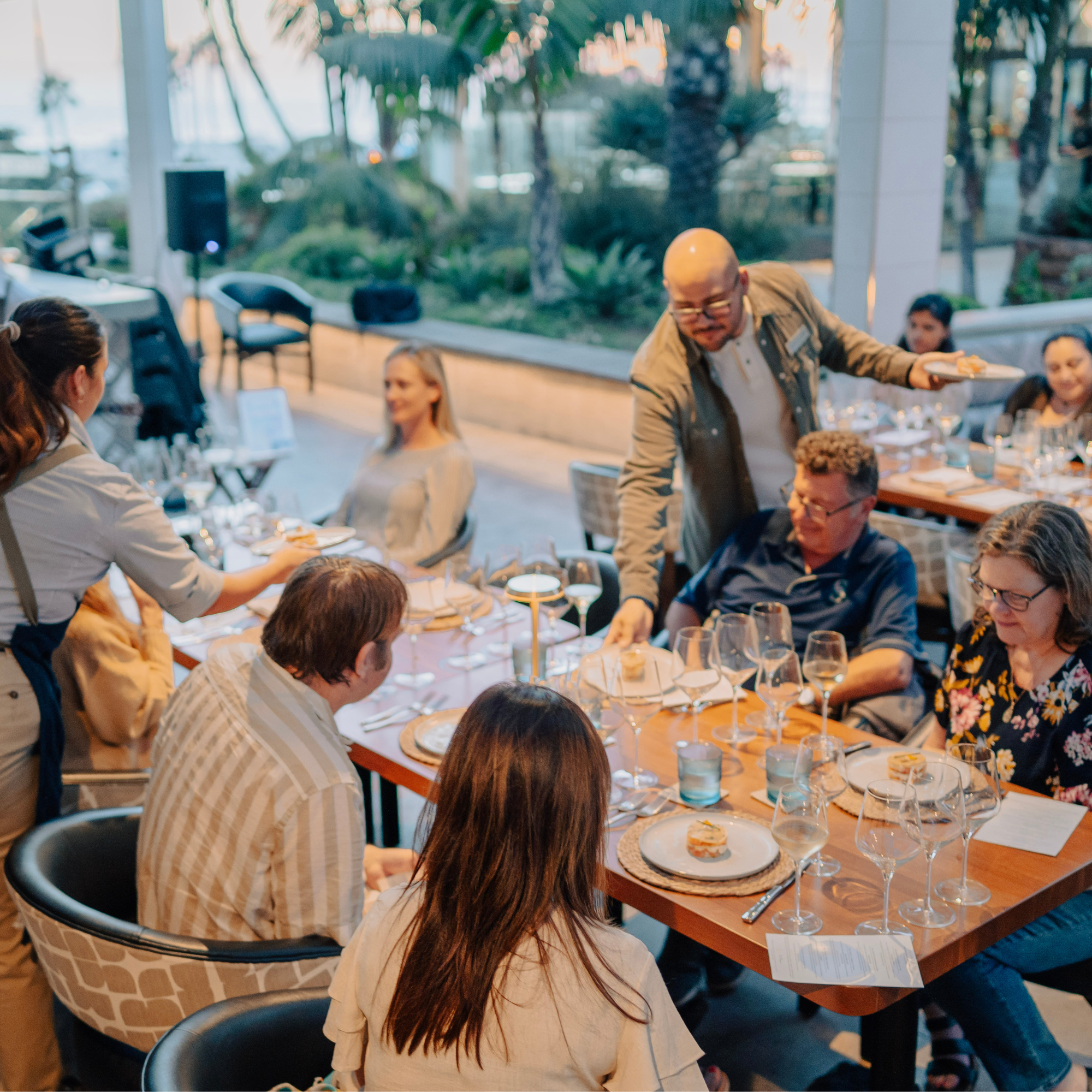 A group of people sit around a restaurant table outdoors, engaging in conversation while staff serve food and set the table.