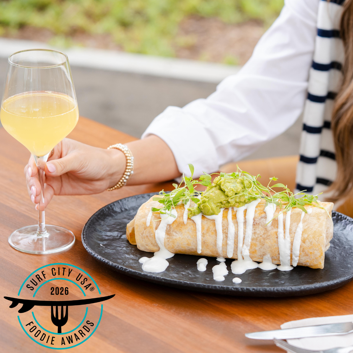 A person holds a glass of white wine next to a large burrito topped with sauce, guacamole, and microgreens on a black plate. A "Surf City USA Foodie Awards 2026" badge is visible.