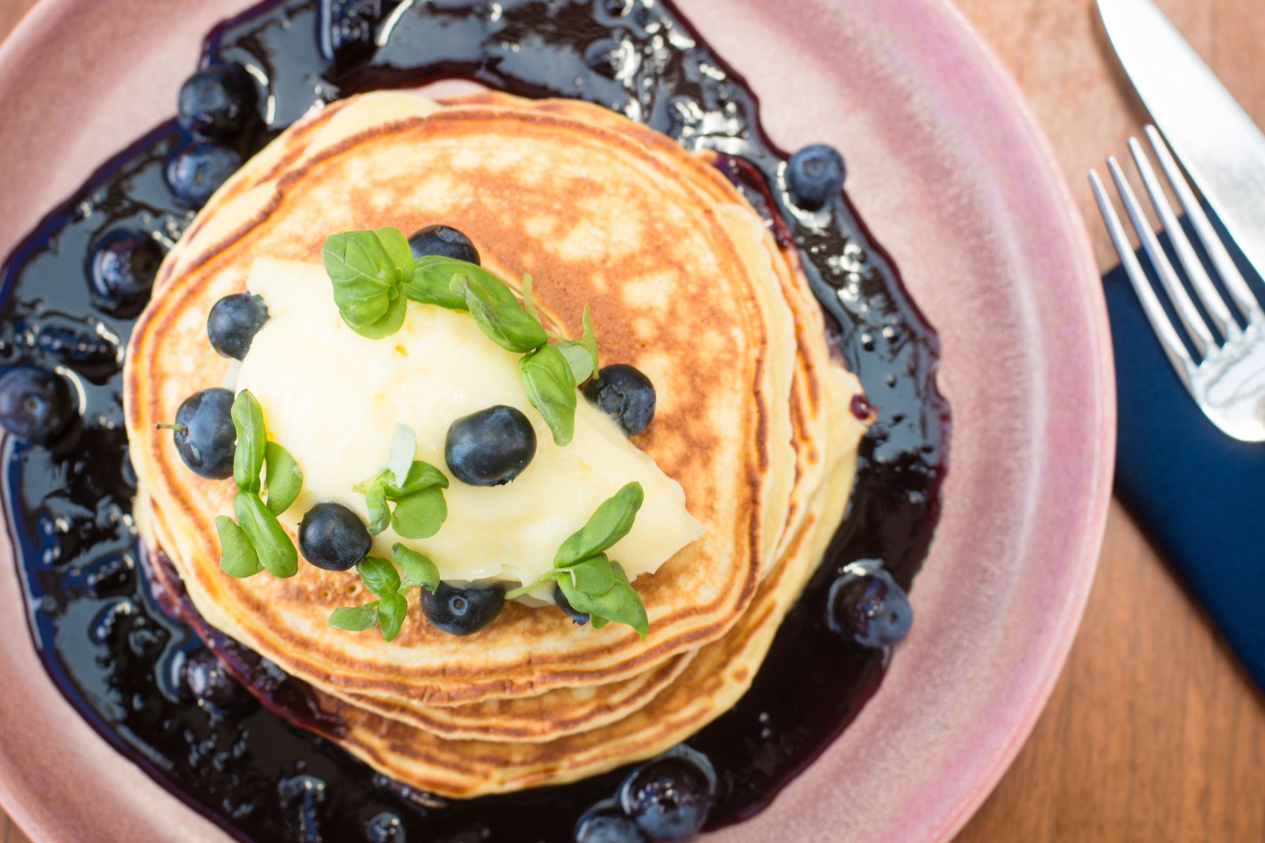 A stack of pancakes topped with butter, blueberries, and greens, served with blueberry sauce on a pink plate—perfect for a Father's Day breakfast at Lōrea, with a fork and knife ready to enjoy.