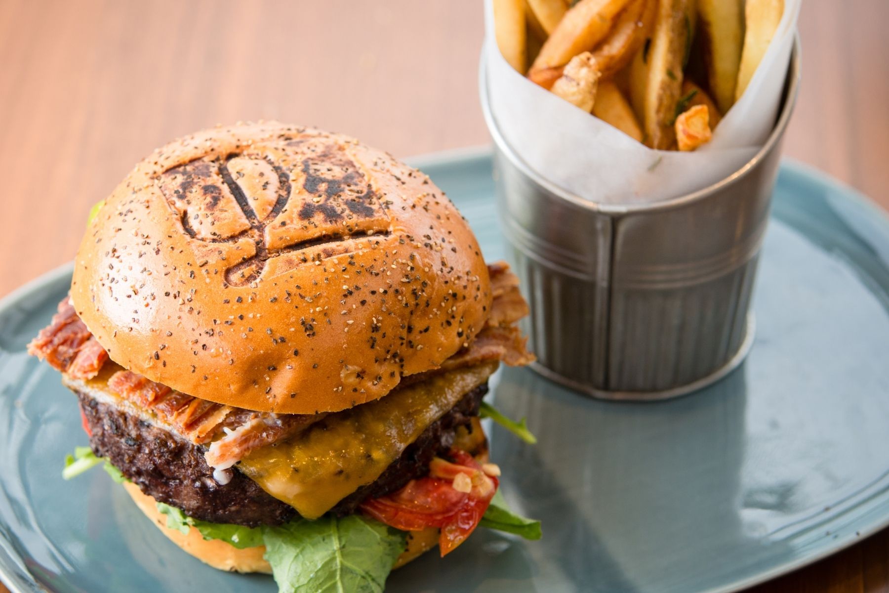 A cheeseburger with lettuce, tomato, and bacon on a seeded bun is served with a tin of fries on a blue plate—perfect for celebrating Burger Week in Orange County.
