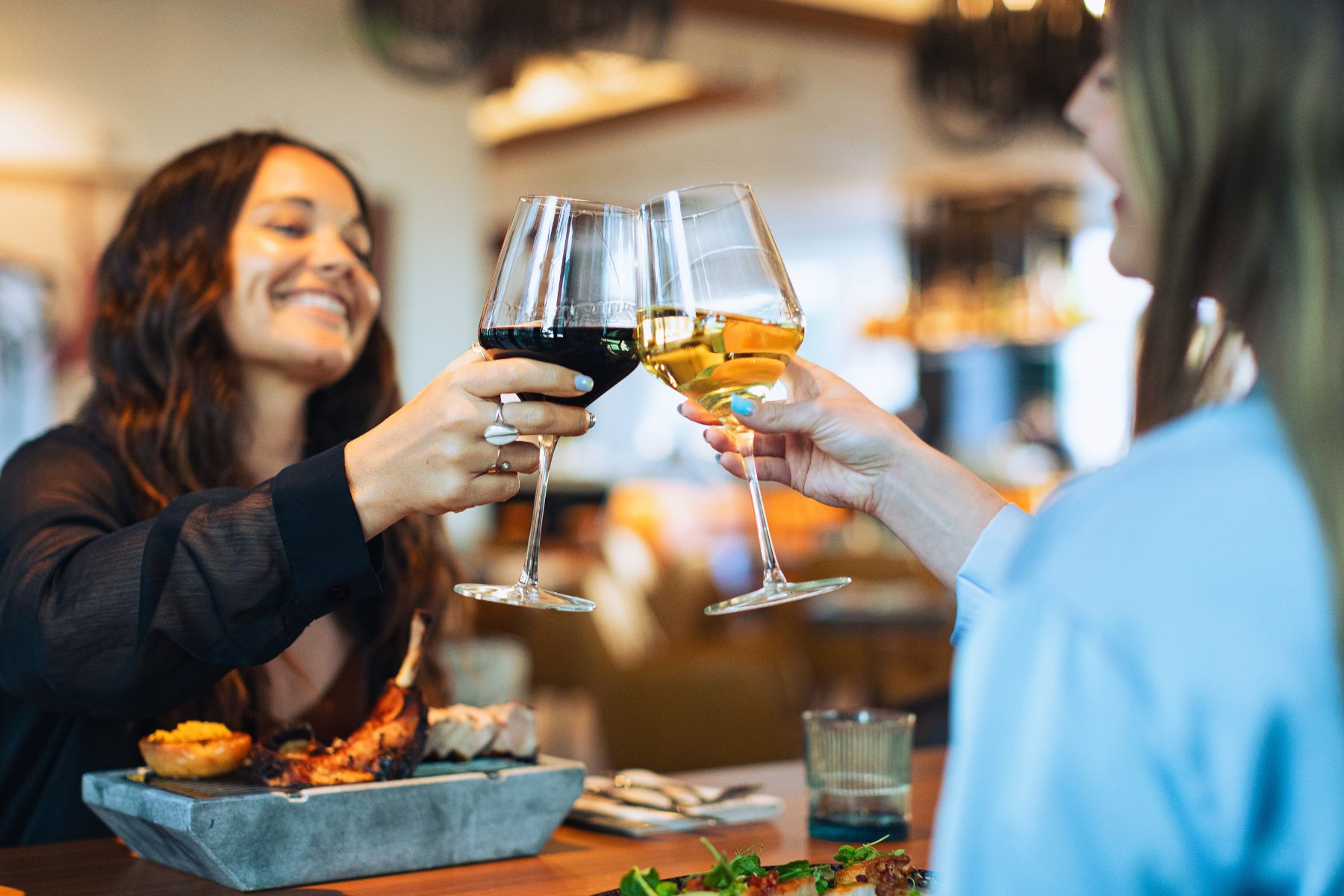 Two people clink wine glasses across a dining table in a restaurant, enjoying plates of food and drinks in front of them—a perfect kickoff dinner to celebrate the upcoming Food Festival.